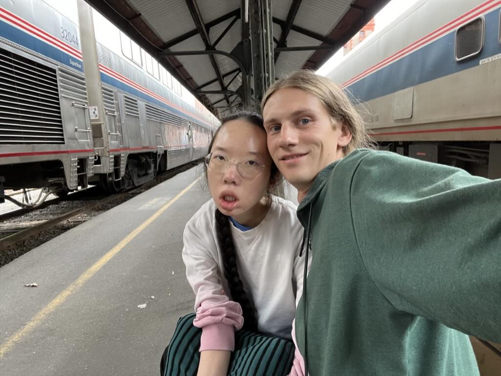 A selfie taken by Peter of him and Aubrie sitting on a station bench during an Amtrak stop in Portland. Superliner cars sit on both sides of the center island platform.