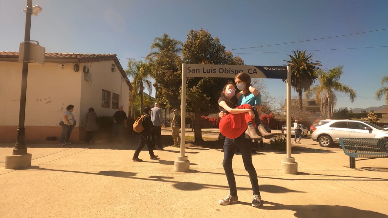 View out the train window of Peter carrying Aubrie around the San Luis Obispo station. Other passengers walk and wait around in the background.
