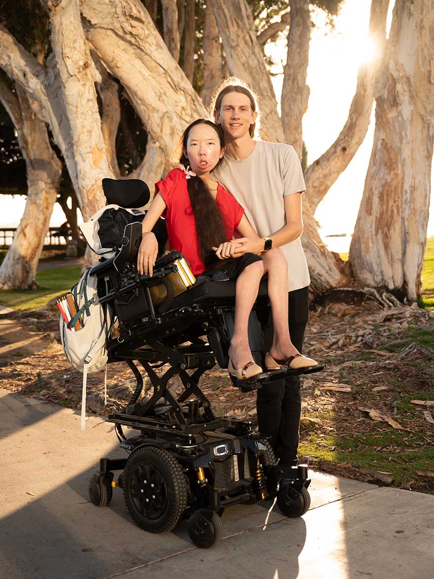 Aubrie and Peter at sunset in front of giant eucalyptus trees. Peter's backpack holds his folded white cane, Aubrie's wheelchair holds Peter's backpack, and Aubrie and Peter hold hands.